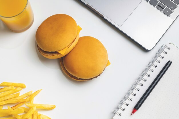 Comida rápida en el trabajo de merienda. Computadora portátil, dos hamburguesas y papas fritas en el lugar de trabajo.