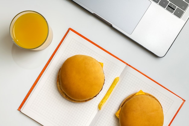 Comida rápida en el trabajo de merienda. Ordenador portátil, cuaderno, dos hamburguesas y papas fritas en el lugar de trabajo.