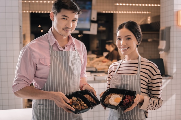 Comida saborosa. Dois garçons simpáticos e radiantes segurando belas lancheiras com comida saborosa parados perto da cozinha