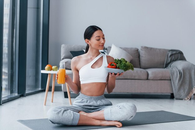 Con comida saludable y jugo de naranja en las manos Mujer joven con forma de cuerpo delgado en ropa deportiva tiene un día de fitness en casa