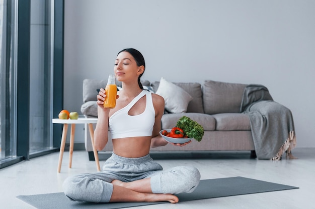 Con comida saludable y jugo de naranja en las manos Mujer joven con forma de cuerpo delgado en ropa deportiva tiene un día de fitness en casa