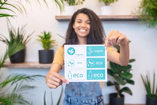 Comida saludable. Mujer feliz mostrando carteles con símbolos de productos útiles en manos extendidas de pie cerca de plantas de interior