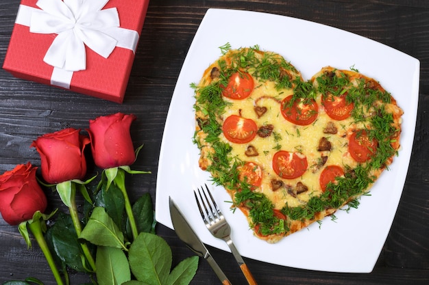 Comida de San Valentín. Pizza en forma de corazón junto a un regalo y rosas rojas, sobre un fondo de madera oscura.