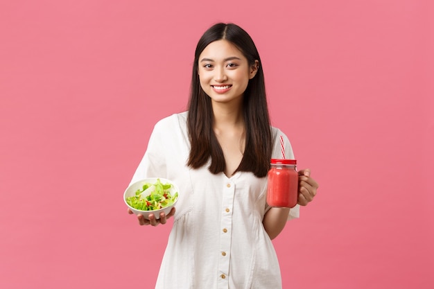 Comida sana, emociones y concepto de estilo de vida de verano. Atractiva mujer joven de 20 años cuidando el cuerpo, comiendo ensalada y bebiendo batidos frescos, de pie con fondo rosa.