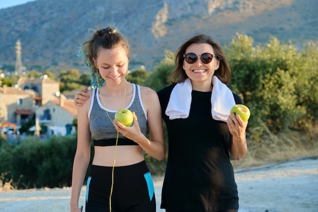 Comida sana, estilo de vida activo. Madre e hija adolescente comiendo manzanas en ropa de fitness después de trotar en una carretera de montaña al atardecer