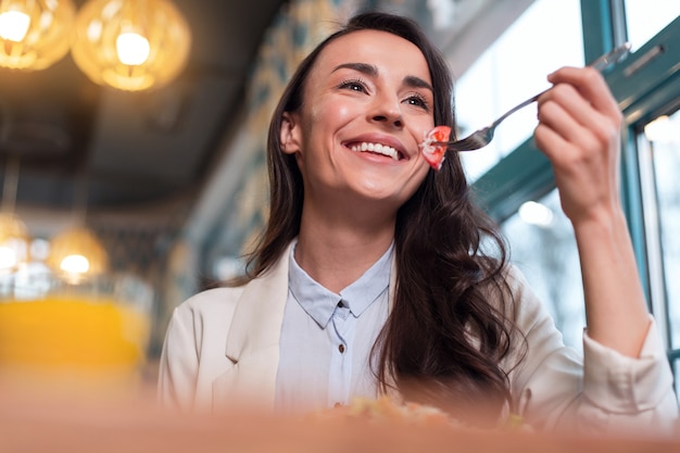 Comida sana. Ángulo bajo de mujer alegre bastante optimista sosteniendo un tenedor mientras se ríe y come tomate