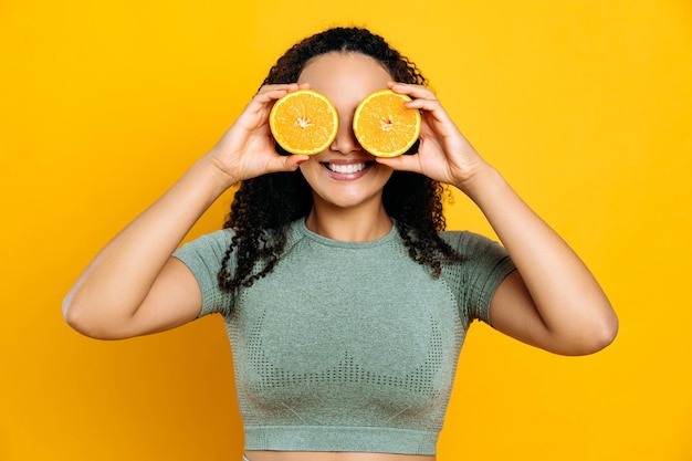 Comida saudável e desintoxicação do estilo de vida Alegre linda raça mista jovem mulher de cabelos cacheados em roupas esportivas em pé no fundo laranja isolado tem duas metades laranja nas mãos perto dos olhos sorrindo