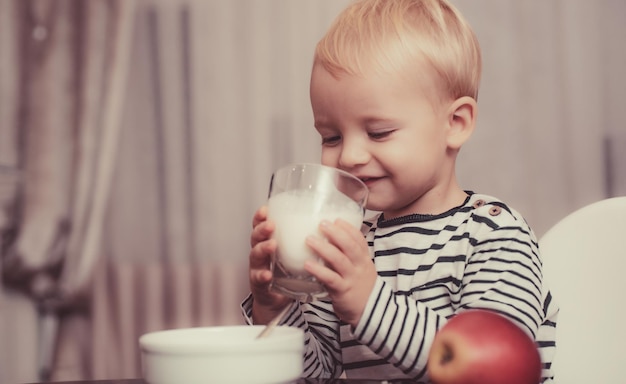 Comida saudável Menino bebê fofo tomando café da manhã Nutrição do bebê Coma saudável Criança fazendo lanche Nutrição saudável Beba leite Criança segura copo de leite Garoto menino fofo sentado à mesa com prato e comida