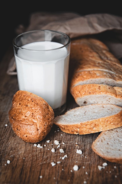 Comida saudável. Um longo pedaço de pão rural com duas peças cortadas repousa sobre uma tábua de cortar de madeira e um copo de leite fresco. Fundo escuro.