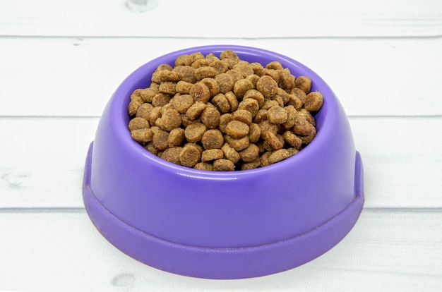 Comida seca para perros en un recipiente de plástico púrpura sobre un piso de madera clara.