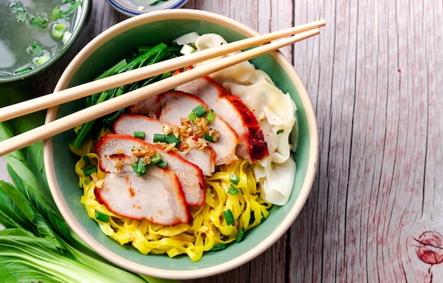 Foto comida tailandesa: fideos de huevo (amarillo) con cerdo rojo asado en un tazón verde y un palillo encima del que se coloca en la mesa de madera en la vista superior