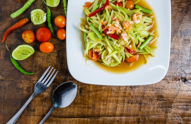 Comida tailandesa, salada de papaia e tomate, pimentão e condimentos em uma mesa de madeira