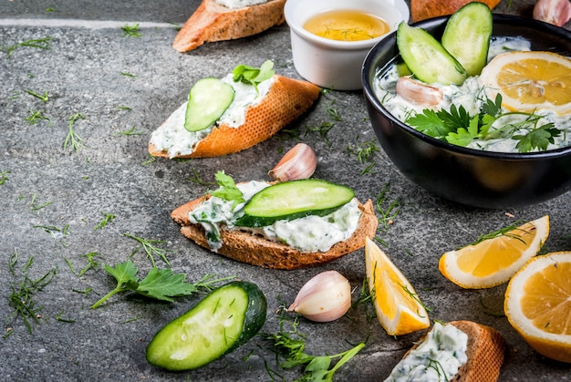 Comida tradicional caucasiana e grega. Molho tzatziki com ingredientes - pepino, limão, salsa, endro, alho. Em uma mesa de pedra escura. Com sanduíches e baguete. Copie o espaço