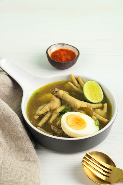 Comida tradicional indonesia llamada Soto Ceker Ayam (sopa de pollo con pierna de pollo) en un tazón de cerámica marrón, con sambal, huevo cocido y lima