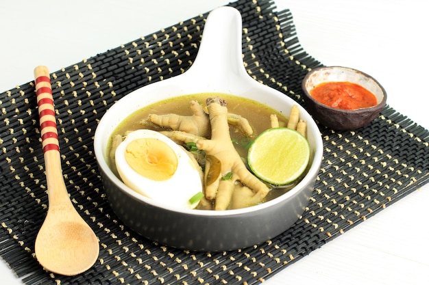 Comida tradicional indonesia llamada Soto Ceker Ayam (sopa de pollo con pierna de pollo) en un tazón de cerámica marrón, con sambal, huevo cocido y lima