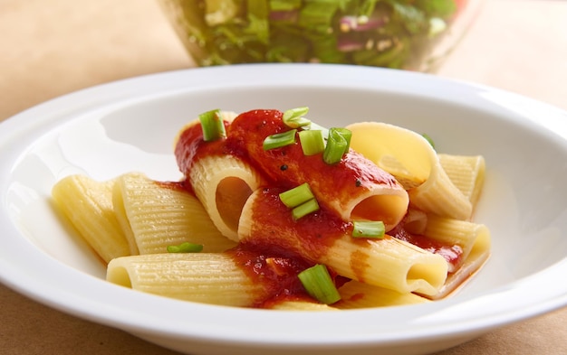 Comida vegana saludable de una pasta italiana cocida tradicional con salsa de tomate y guarnición de cebolla verde contra el tazón borroso recortado con ensalada de hierbas saludables