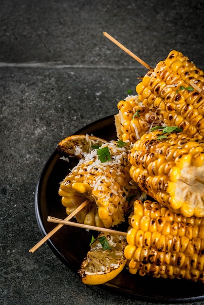 Comida de verano Ideas para fiestas de barbacoa y parrilla. Maíz a la parrilla a la parrilla en llamas. Con una pizca de queso (elotes mexicanos), ají picante, limón. Sobre una mesa de piedra oscura, placa negra Copia espacio