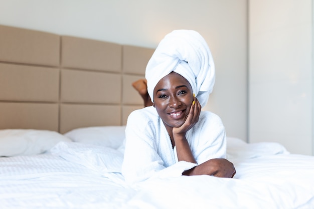 Comienzo positivo del día. Mujer joven africana sonriente que miente en cama en albornoz.
