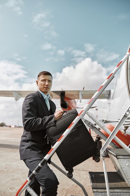 Comissária de bordo subindo com a bagagem na escada para o jato do avião. Avião de passageiros moderno. Homem sério multirracial usa uniforme. Dia ensolarado. Aviação comercial civil. Conceito de viagem aérea