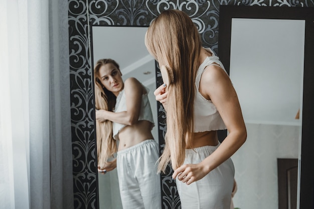 Foto cómo cuidar el cabello largo vista posterior de la joven rubia natural en ropa blanca para el hogar peinándola