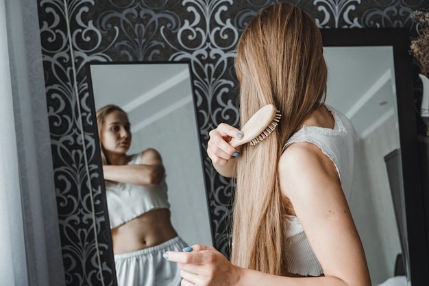 Cómo cuidar el cabello largo vista posterior de la joven rubia natural en ropa blanca para el hogar peinándola