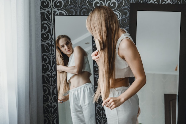 Foto como cuidar do cabelo comprido, vista traseira de uma jovem mulher loira natural com roupas brancas em casa penteando-a