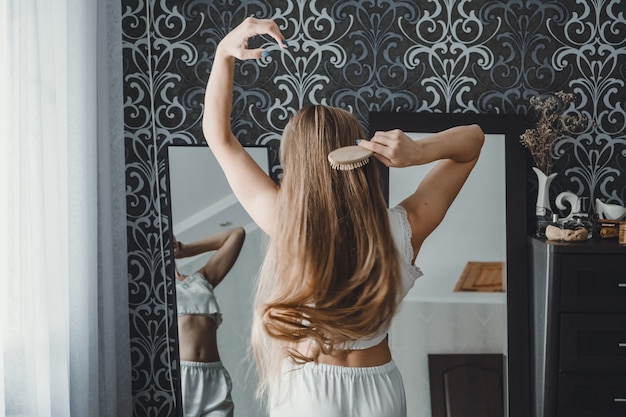 Como cuidar do cabelo comprido, vista traseira de uma jovem mulher loira natural com roupas brancas em casa penteando-a