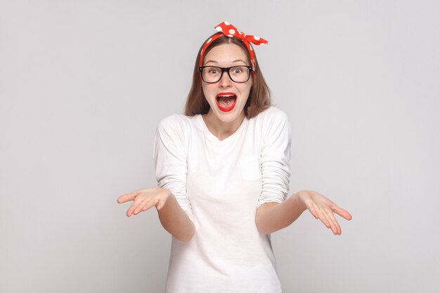 ¿Cómo hiciste esto? es increíble. Retrato de hermosa mujer joven emocional en camiseta blanca con pecas, gafas negras, labios rojos y banda para la cabeza. tiro interior, aislado sobre fondo gris claro.