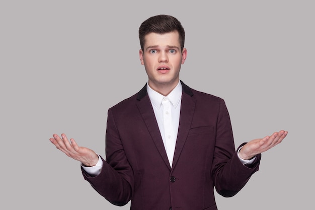 ¿Cómo hiciste esto? Retrato de confundido joven guapo con traje violeta y camisa blanca, de pie, mirando a cámara con los brazos levantados y preguntando. tiro del estudio de interior, aislado en fondo gris.