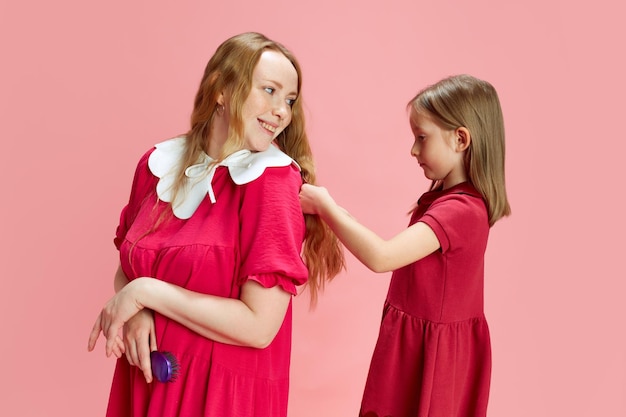 Como mamá como hija, niña peinándose a su madre contra el fondo rosa del estudio