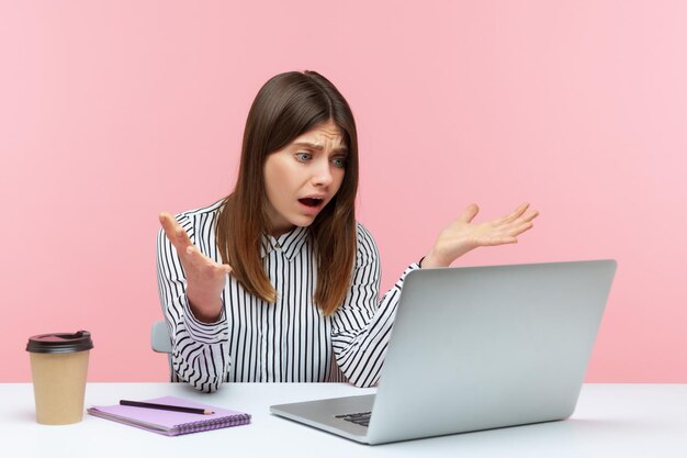 Como pudiste. Trabajadora de oficina irritada levantando la mano con un gesto indignado, preguntando por qué y mirando la pantalla del portátil, hablando por videollamada. Disparo de estudio interior aislado sobre fondo rosa