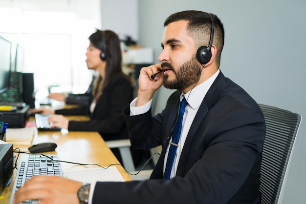 ¿Como puedo ayudarte? Guapo representante de ventas hispano con auriculares hablando con un cliente en un centro de llamadas y una empresa comercial