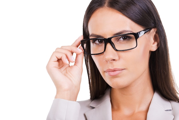 ¿Como puedo ayudarte? Hermosa joven empresaria en traje ajustando sus anteojos y mirando a la cámara mientras está de pie contra el fondo blanco.