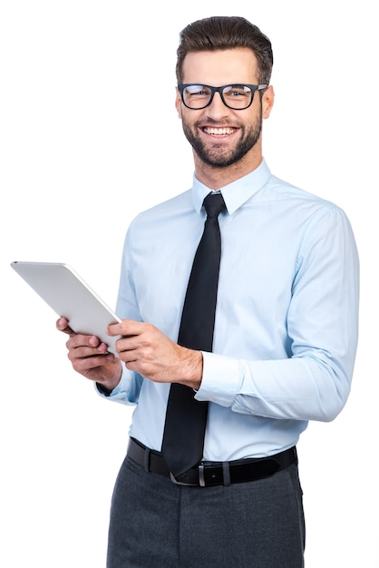 ¿Como puedo ayudarte? Hombre guapo joven confidente en camisa y corbata sosteniendo tableta digital y mirando a cámara con una sonrisa mientras está de pie contra el fondo blanco