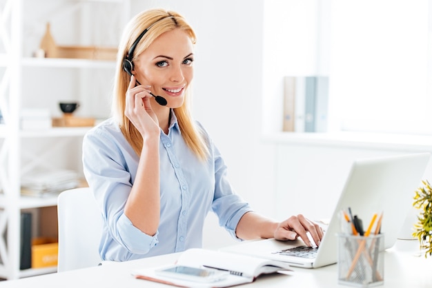 ¿Como puedo ayudarte? Mujer hermosa joven alegre en auriculares trabajando en la computadora portátil y sonriendo mientras está sentado en su lugar de trabajo