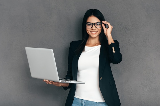 ¿Como puedo ayudarte? Mujer joven atractiva en ropa de sport elegante y anteojos que llevan la computadora portátil y que sonríe mientras está de pie contra el fondo gris