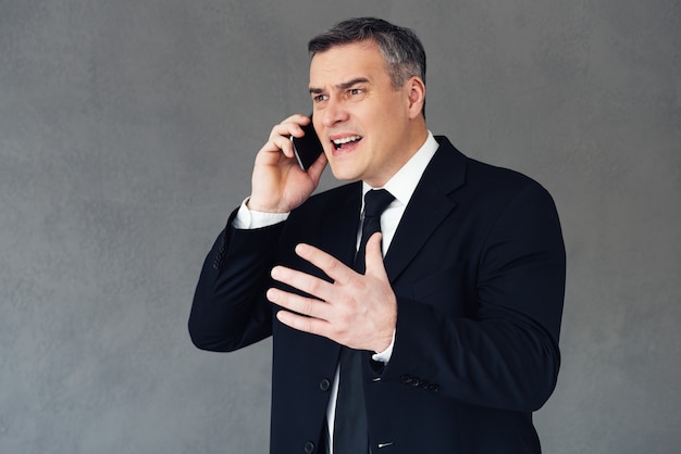 Como você pôde cometer esse erro? Homem de negócios maduro furioso falando no celular e gesticulando em pé contra um fundo cinza
