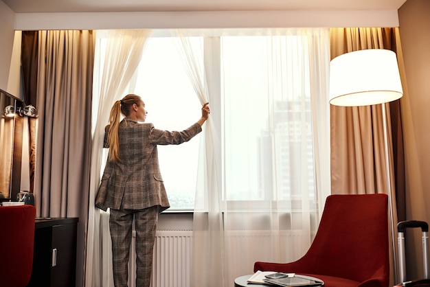 Cómoda habitación de hotel. La empresaria en la oscuridad de la habitación del hotel abre las cortinas de la ventana a la luz de la mañana