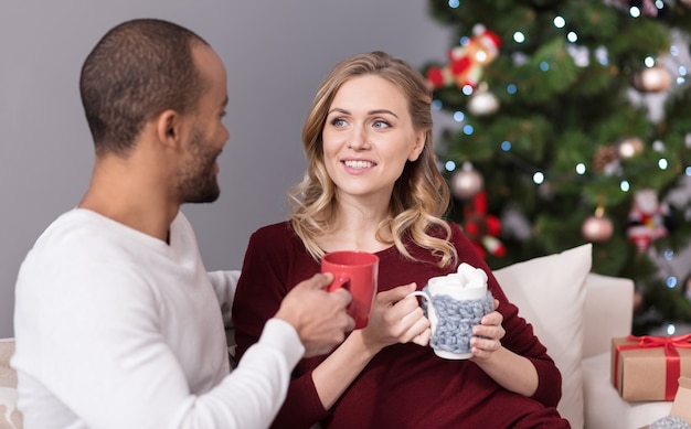 Comodidad hogareña. Feliz pareja encantada agradable sentado en el sofá y sosteniendo tazas mientras toma el té