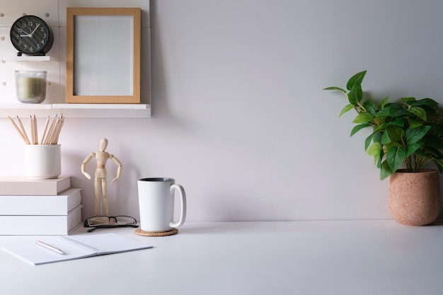 Cómodo lugar de trabajo con marco de imagen de planta de interior, taza de café y artículos de papelería en mesa blanca