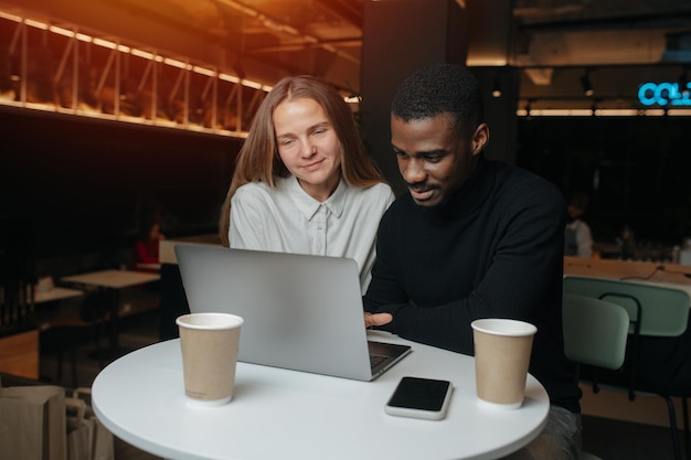 Cómodos amigos interraciales del sexo opuesto sentados en el café, mirando una computadora portátil. A la vez atentos y felices.