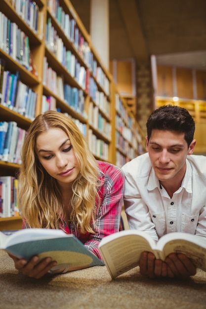 Compañeros de clase sonrientes que leen el libro mientras que se apoyan en estantes en biblioteca