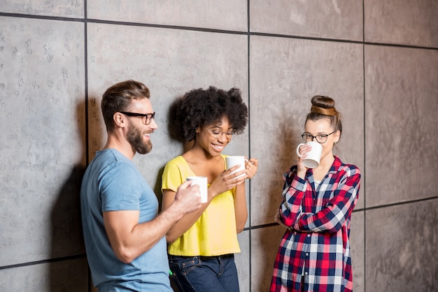 Compañeros de trabajo multiétnicos vestidos casualmente tomando un café cerca de la pared gris en el interior