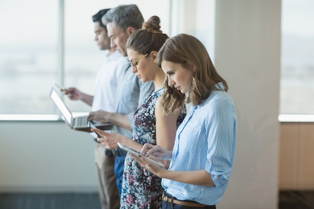 Compañeros de trabajo de pie en una fila con teléfono móvil, tableta digital y computadora portátil