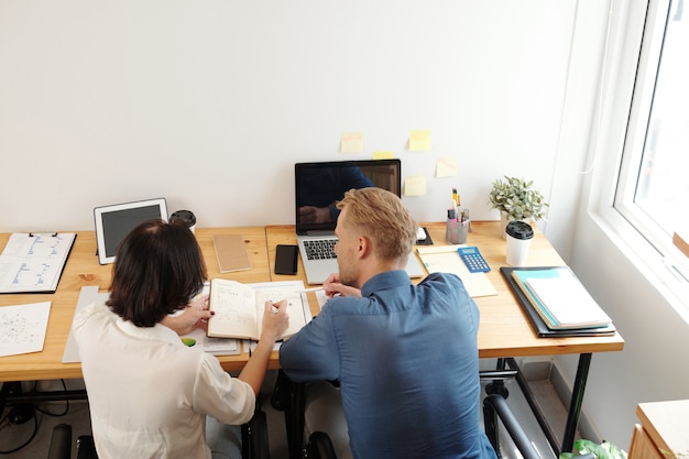 Compañeros de trabajo que escriben en el planificador cuando discuten nuevas ideas y estrategias de desarrollo de la empresa cuando la pandemia ha terminado, vista desde atrás