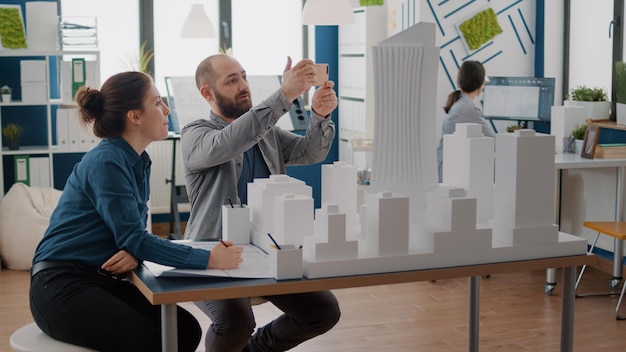 Compañeros de trabajo que usan teléfonos móviles para diseñar el diseño de la construcción después de construir el modelo en la mesa. Hombre y mujer trabajando juntos con un smartphone para planificar un proyecto urbano con maqueta y boceto.