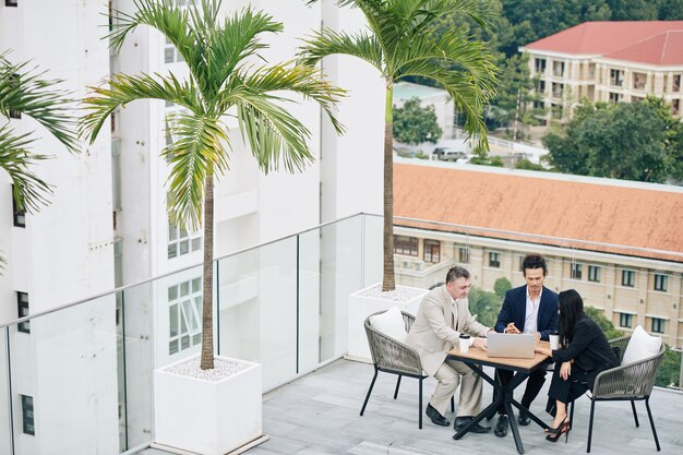 Compañeros de trabajo reunidos en una mesa de café en la azotea y discutiendo datos en la pantalla del portátil