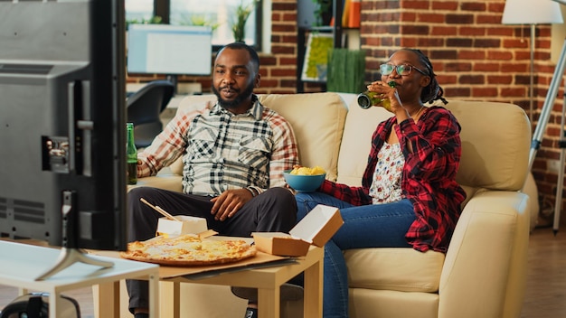 Los compañeros de vida felices se dan un atracón viendo programas de televisión juntos, sosteniendo una botella de cerveza y comiendo papas fritas en casa. Los jóvenes en relación se sienten relajados con una película en la televisión, comiendo bocadillos.