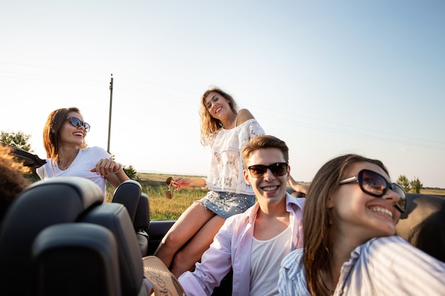 Companhia de garotas e caras estão sentados e sorrindo em um cabriolet preto na estrada rural em um dia ensolarado. .