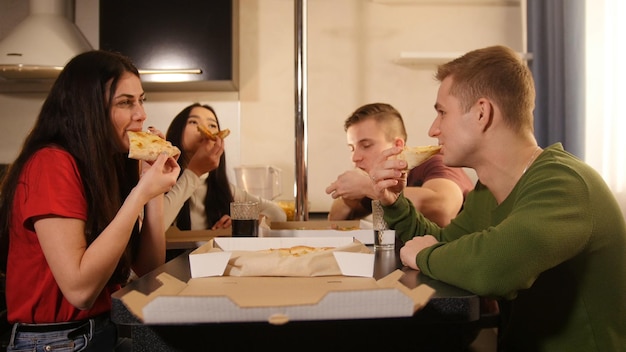 Companhia de jovens amigos sentados na cozinha e comendo pizza da caixa de entrega